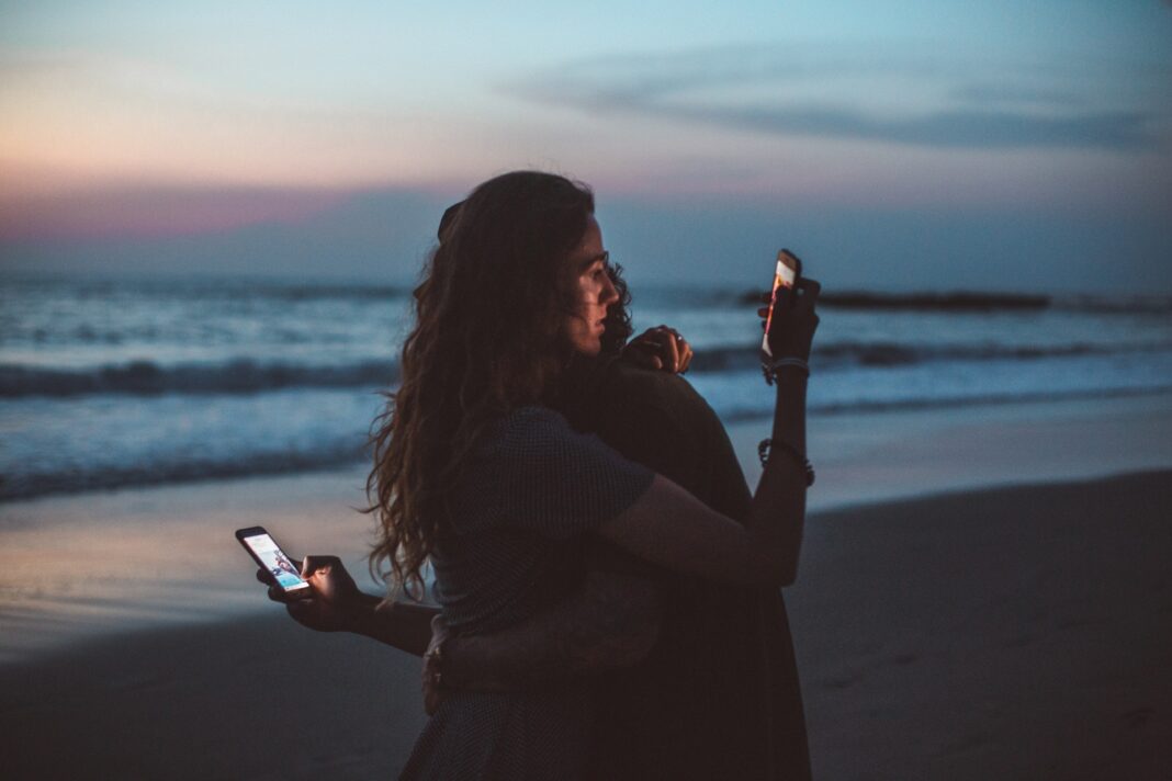 Milenci se objímají na mořském břehu a přitom koukají na svoje mobilní telefony - jak zjistit, komu patří telefonní číslo
